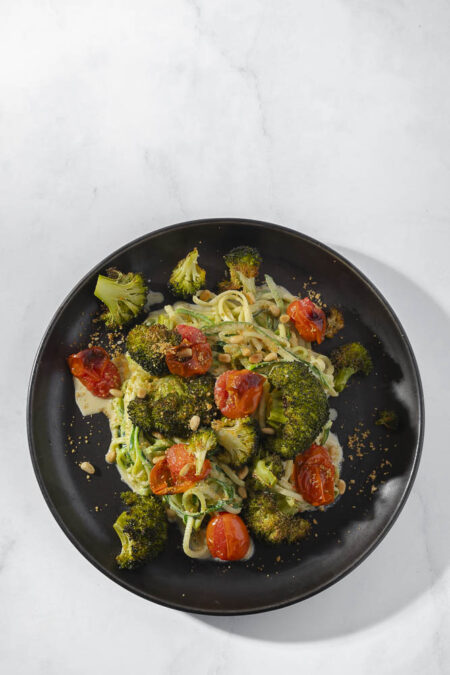 Bovenaanzicht van zwart bord op witte marmeren ondergrond. Op het bord Romige courgettespaghetti met cashew roomsaus en geroosterde broccoli en tomaat met pijnboompitten.