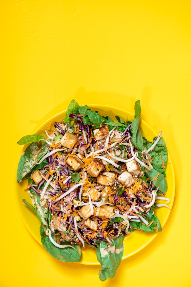 Geel bord met daarop een salade met tofu en pinda dressing, tegen een gele achtergrond